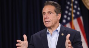 governor cuomo speaking with hands raises to shoulder level. blue sportcoat with no tie, American flag in background